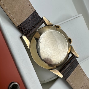 Puede incluir: Un reloj de pulsera dorado con una correa de cuero marr&oacute;n. La esfera del reloj no es visible, pero la parte posterior de la caja del reloj es visible y dice "10K Gold Filled" y "LeCoultre".