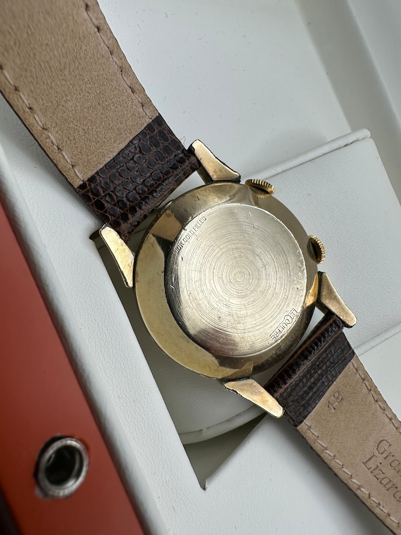 Puede incluir: Un reloj de pulsera dorado con una correa de cuero marr&oacute;n. La esfera del reloj no es visible, pero la parte posterior de la caja del reloj es visible y dice "10K Gold Filled" y "LeCoultre".