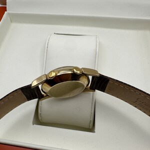 Puede incluir: Un reloj de pulsera dorado con una correa de cuero marr&oacute;n se muestra en una caja blanca con un forro rojo. El reloj tiene una esfera redonda y tres botones en el lateral.