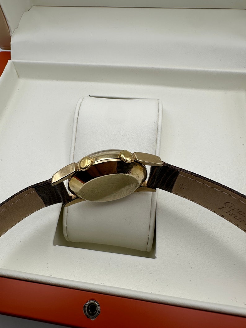 Puede incluir: Un reloj de pulsera dorado con una correa de cuero marr&oacute;n se muestra en una caja blanca con un forro rojo. El reloj tiene una esfera redonda y tres botones en el lateral.