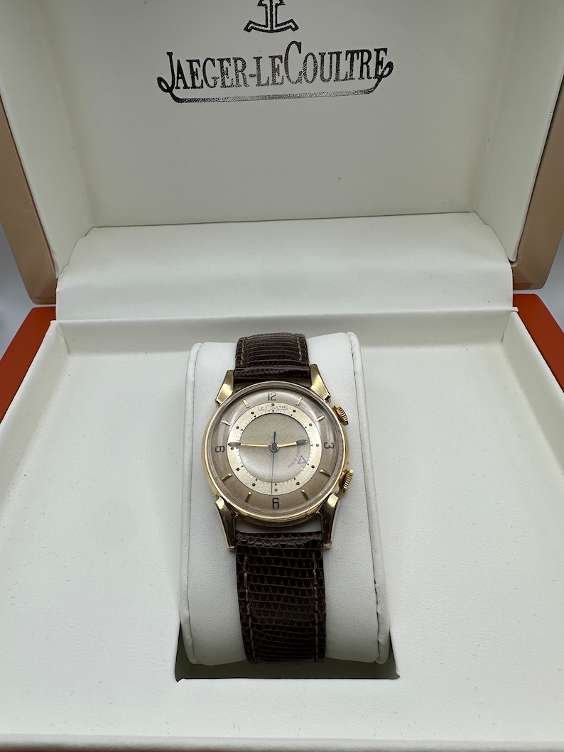 Puede incluir: Un reloj de pulsera dorado con una correa de cuero marr&oacute;n, en una caja blanca con el texto "JAEGER-LECOULTRE" en la tapa.