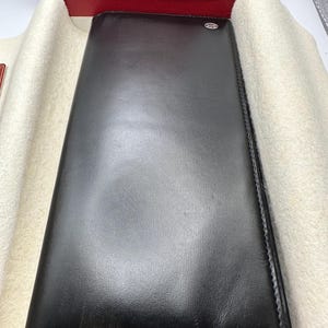 May include: A black leather long wallet is displayed in a red box. The wallet has a subtle logo and is neatly stitched.  The smooth leather exterior shows minimal texture. The wallet is presented on a cream-colored fabric lining within the box. This luxury leather wallet is ideal for storing cards, cash, and other essentials.