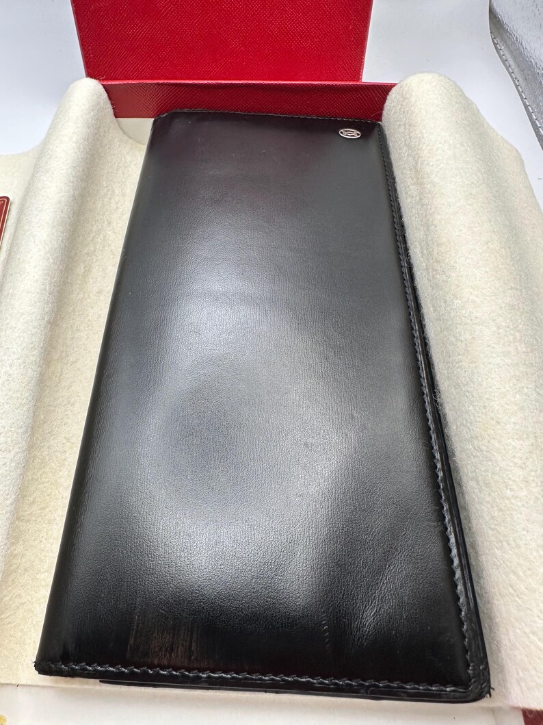 May include: A black leather long wallet is displayed in a red box. The wallet has a subtle logo and is neatly stitched.  The smooth leather exterior shows minimal texture. The wallet is presented on a cream-colored fabric lining within the box. This luxury leather wallet is ideal for storing cards, cash, and other essentials.