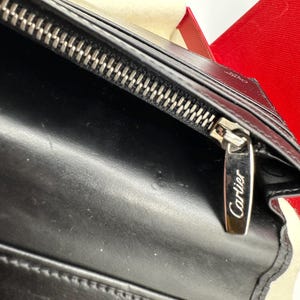 May include: Close-up of a black leather Cartier wallet. The wallet features a silver zipper with the Cartier logo engraved on the zipper pull. The leather appears smooth and luxurious. The wallet is partially shown in a red box.