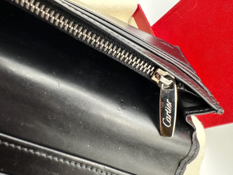 May include: Close-up of a black leather Cartier wallet. The wallet features a silver zipper with the Cartier logo engraved on the zipper pull. The leather appears smooth and luxurious. The wallet is partially shown in a red box.
