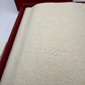 May include: Close-up view of a cream-colored, felt-like Cartier jewelry box interior. The box is lined with a soft, textured fabric. The Cartier logo is subtly embossed in the center. The overall color palette is neutral and elegant, suggesting high-end packaging for luxury goods.