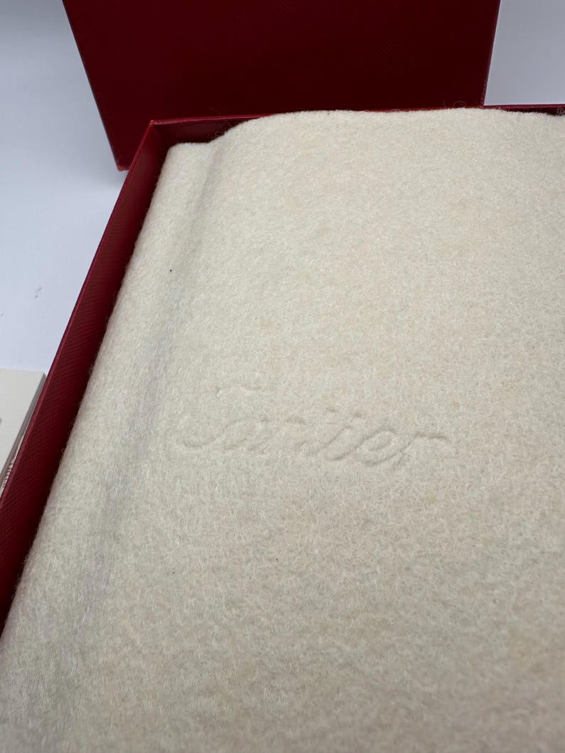 May include: Close-up view of a cream-colored, felt-like Cartier jewelry box interior. The box is lined with a soft, textured fabric. The Cartier logo is subtly embossed in the center. The overall color palette is neutral and elegant, suggesting high-end packaging for luxury goods.