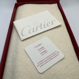 May include: Cartier leather goods packaging.  A red box contains a beige felt insert.  Two white cards display the Cartier logo and product details: exterior and interior are calfskin leather.  Care instructions advise handling with care and avoiding humidity.