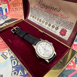 Puede incluir: Un reloj de pulsera Longines de oro con una esfera blanca y una correa de cuero negro, presentado en una caja de terciopelo rojo con ribete dorado. La tapa de la caja presenta una inscripción dorada que dice "Longines The World's Most Honored Watch".