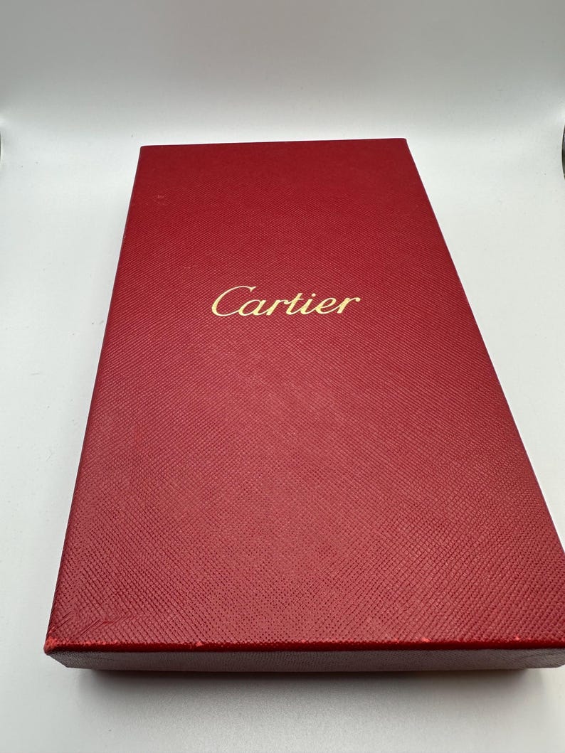 May include: A red Cartier box. The box is rectangular and made of a textured material. The Cartier logo is embossed in gold on the front of the box. The box is a deep, rich red color.  The box is likely used for packaging luxury goods. The box is shown from a slightly elevated angle, showcasing its top surface and sides.