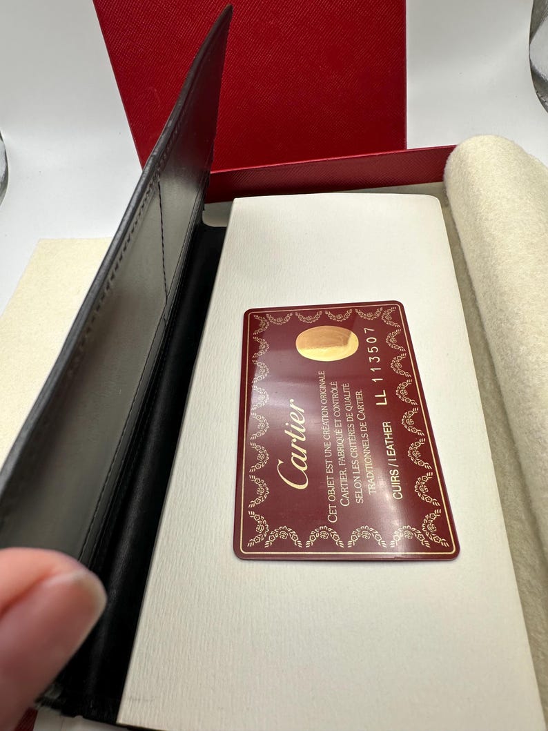 May include: Close-up view of a black leather Cartier pen case, partially open, revealing a white interior and a burgundy Cartier authenticity card. The card displays the Cartier logo and text indicating the item is an original creation, made and controlled according to Cartier's quality criteria, and specifies the leather type and a product code: LL 113507. The case shows fine stitching and a sleek design, typical of luxury goods.
