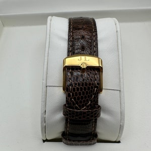 Puede incluir: Una correa de reloj de cuero marr&oacute;n con una hebilla dorada que dice "JL".