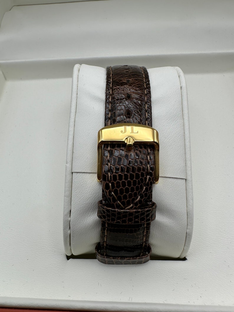 Puede incluir: Una correa de reloj de cuero marr&oacute;n con una hebilla dorada que dice "JL".