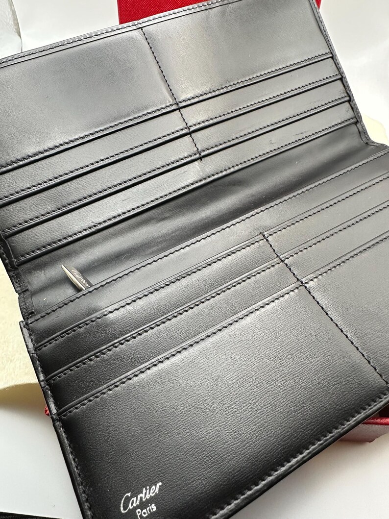May include: A black leather Cartier Paris wallet is open, showcasing multiple card slots and compartments.  The interior is neatly stitched, and the brand name is subtly embossed on the bottom.  The wallet appears to be made of high-quality leather with a smooth finish. A silver pen is partially visible in one of the compartments.