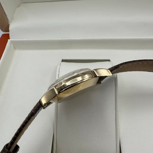 Puede incluir: Un reloj de pulsera dorado con una correa de cuero marr&oacute;n, presentado en una caja blanca.