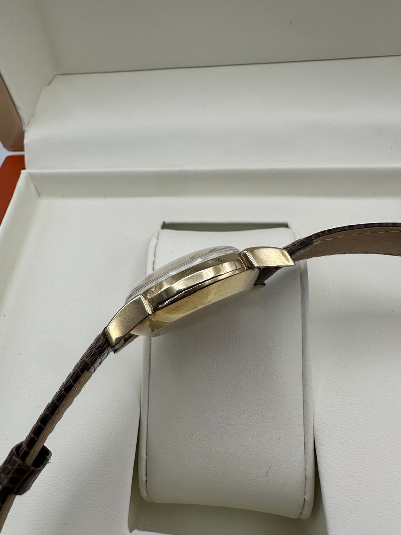 Puede incluir: Un reloj de pulsera dorado con una correa de cuero marr&oacute;n, presentado en una caja blanca.