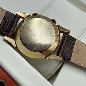 Puede incluir: Un reloj de pulsera dorado con una correa de cuero marr&oacute;n. La parte posterior del reloj tiene el texto "LeCoultre" grabado.