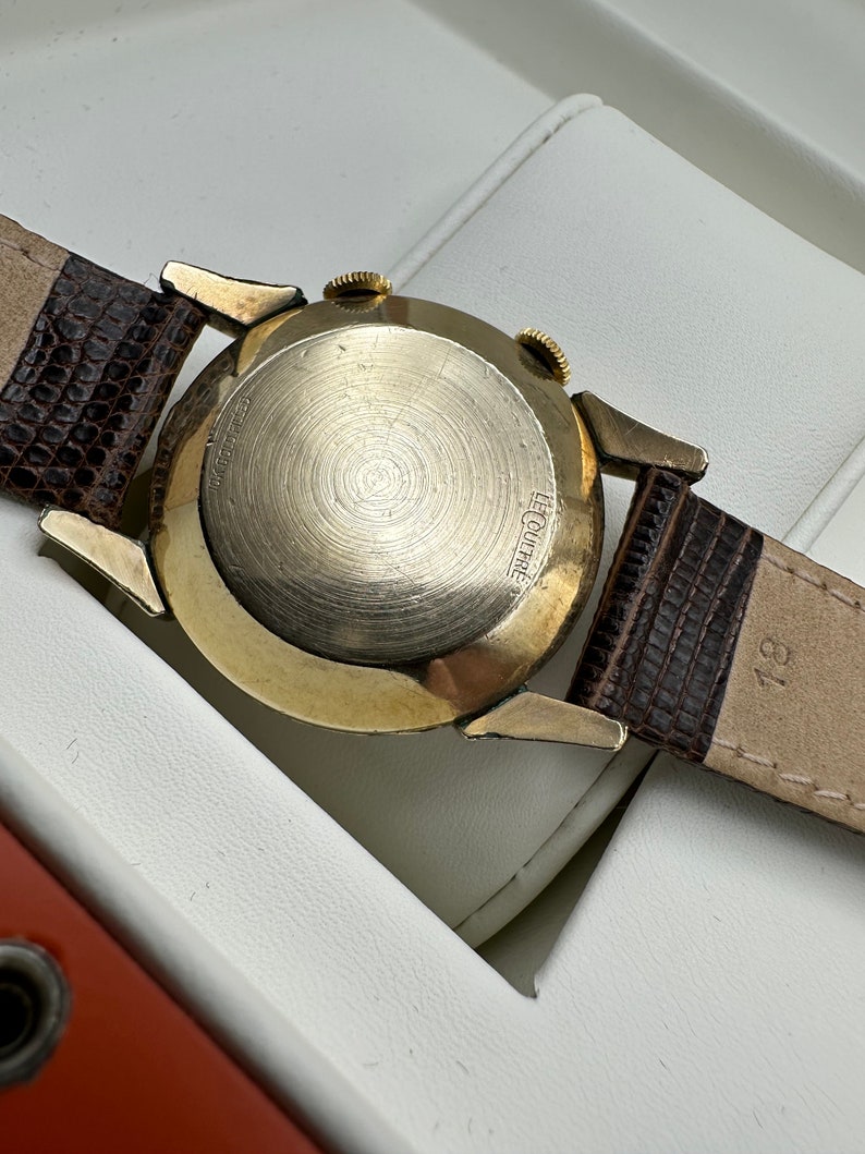 Puede incluir: Un reloj de pulsera dorado con una correa de cuero marr&oacute;n. La parte posterior del reloj tiene el texto "LeCoultre" grabado.