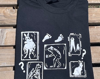 May include: Black t-shirt with a white graphic print of a series of framed images. The images include a moth, footprints, a frog, a pair of figures, a rabbit, and a creature with wings. The text "?" appears twice on the shirt.