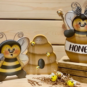 May include: Three wooden bee figurines, one holding a jar of honey with the word "Honey" written on it, another standing on its own, and a third beehive with bees crawling around it.