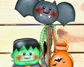 May include: A Halloween wooden decoration with a bat, Frankenstein's monster, and a potion bottle. The bat is grey with a smiling face. The monster is green with black hair. The potion bottle is orange with bats. The base reads "PICK YOUR POISON".