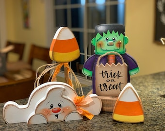May include: Halloween decorations featuring a white ghost, a green Frankenstein, and two candy corn figures. The Frankenstein figure is holding a brown paper bag with the words "trick or treat" written on it.