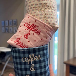 May include: Three fabric coffee cup sleeves stacked. The top sleeve is cream with a floral pattern and the words "coffee & Jesus" embroidered in beige. The middle sleeve is pink with red hearts and the same words embroidered in red. The bottom sleeve is blue and black checkered with the same words embroidered in beige.