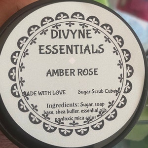 May include: A white jar with a black lid and a label that says "DIVYNE ESSENTIALS AMBER ROSE" and "MADE WITH LOVE Sugar Scrub Cubes". The label also lists the ingredients: "Sugar, soap base, shea butter, essential oils, nontoxic mica color".