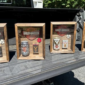 Könnte beinhalten: Vier rechteckige Schattenboxen aus Holz mit dem Harley-Davidson Motor Cycles Logo im oberen Bereich. Jede Vitrine enthält eine Vintage-Getränkedose, eine kleine rechteckige Schachtel und einen runden Chip, die hinter einer Glasscheibe angeordnet sind.