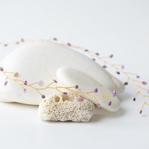 May include: A delicate gold wire headband featuring small, light purple amethyst gemstones. The floral design is displayed on a white background with a natural stone. The gemstones are approximately 0.6 cm in size.