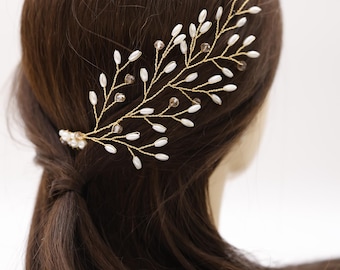 May include: A gold-toned hair accessory shaped like a delicate branch, adorned with white pearl-like beads and small, clear crystals. The hairpiece is set against a backdrop of dark brown hair, suggesting a bridal or special occasion theme.