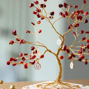 Op de afbeelding: Een gouden draadboom met rode kralen en witte keramische bedels. De bedels zijn hartvormig en hebben de letters "R" en "A" erop.