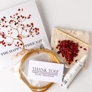 Puede incluir: Un kit de manualidades para hacer un árbol de alambre con cuentas rojas. El kit incluye una tarjeta con las palabras "The Happy Tree Kit", un rollo de alambre dorado, una bolsa de cuentas rojas, un pequeño tubo de pegamento, un palo de madera y una tarjeta con las palabras "¡Gracias!" e información de redes sociales.