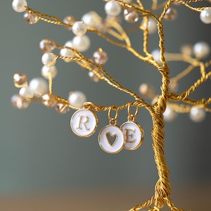 May include: A handcrafted gold-toned wire tree adorned with faux pearls and crystals. The tree features three white enamel charms with the letters "R", "E", and a heart symbol. The tree's branches are embellished with white pearls and amber-colored crystals.