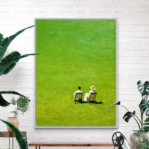 May include: A framed wall art print featuring a vibrant green field with two figures sitting in wooden folding chairs. One figure wears a straw hat and white clothing, while the other wears dark clothing. The scene is minimalist and expansive.
