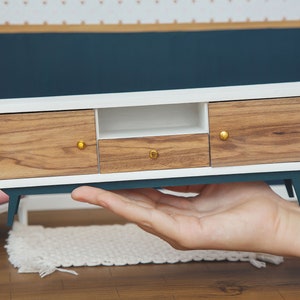 May include: A miniature white and wood media console with gold knobs. The console has two doors, a shelf, and a drawer. The console is sitting on a hand.