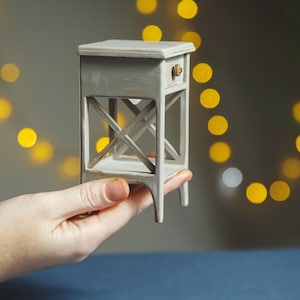 May include: A small, white painted wooden end table with a single drawer and a criss-cross design on the sides. The table is being held in a person's hand.