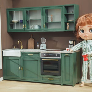 Peut inclure: Une poupée en pyjama rayé vert et blanc se tient dans une cuisine miniature avec des armoires vertes, un évier blanc et un four noir. La poupée tient une tasse rouge.