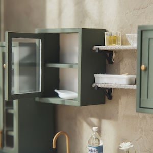 Op de afbeelding: Een miniatuurkeukenset met groene kasten, open planken en glazen deuren. De set bevat een klein wit bord, een witte container en twee glazen potten met een gele vloeistof.