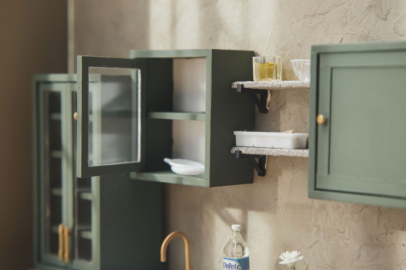 Op de afbeelding: Een miniatuurkeukenset met groene kasten, open planken en glazen deuren. De set bevat een klein wit bord, een witte container en twee glazen potten met een gele vloeistof.