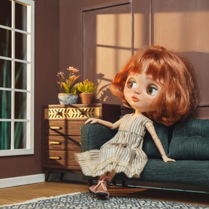 May include: A red-haired doll wearing a striped dress sits on a dark green sofa in a dollhouse setting. The doll has large, blue eyes and is looking to the right. The sofa is in front of a brown wall with a window and a wooden dresser.