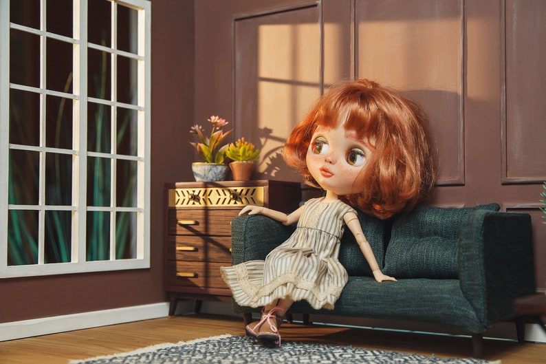 May include: A red-haired doll wearing a striped dress sits on a dark green sofa in a dollhouse setting. The doll has large, blue eyes and is looking to the right. The sofa is in front of a brown wall with a window and a wooden dresser.