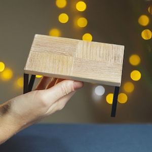 May include: A small wooden table with black metal legs. The table is being held in a hand.