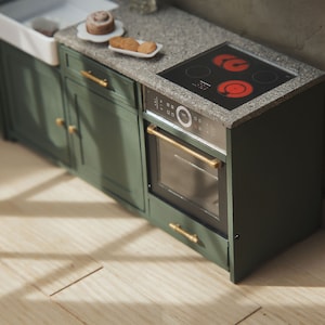 Op de afbeelding: Een groene keukenkast met een zwarte oven en een glazen kookplaat. De kookplaat heeft twee rode hete branders. De kast heeft gouden handgrepen.