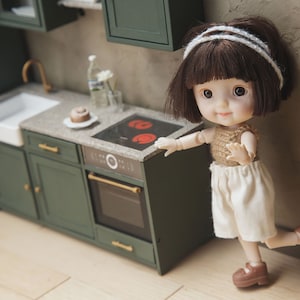 Op de afbeelding: Een pop met bruin haar en een wit hoofdband staat voor een miniatuurkeuken. De keuken heeft groene kasten, een witte gootsteen en een zwart fornuis met een rode brander.