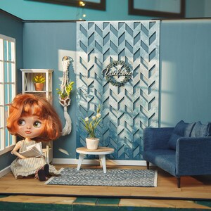 May include: A dollhouse living room with blue walls, a blue couch, a white coffee table, a rug, and a doll sitting on a chair reading a book. The wall has a decorative wooden panel with a geometric pattern and the words "Welcome" on a wreath.