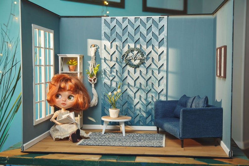 May include: A dollhouse living room with blue walls, a blue couch, a white coffee table, a rug, and a doll sitting on a chair reading a book. The wall has a decorative wooden panel with a geometric pattern and the words "Welcome" on a wreath.
