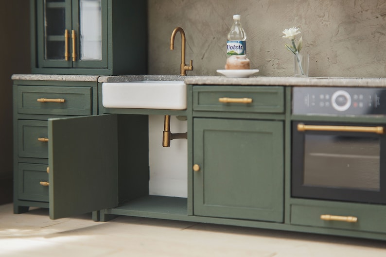 Op de afbeelding: Een miniatuurkeukenset met groene kasten, een witte porseleinen gootsteen, een gouden kraan en een zwarte oven met een gouden handvat. Het aanrecht is grijs en er staat een fles water en een bord met een gebakje op.