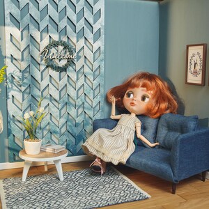 May include: A dollhouse living room with a blue couch, a white bookshelf, a small side table, and a doll with red hair sitting on the couch. The wall is blue and has a decorative wall panel with a geometric pattern. There is a welcome sign hanging on the wall.