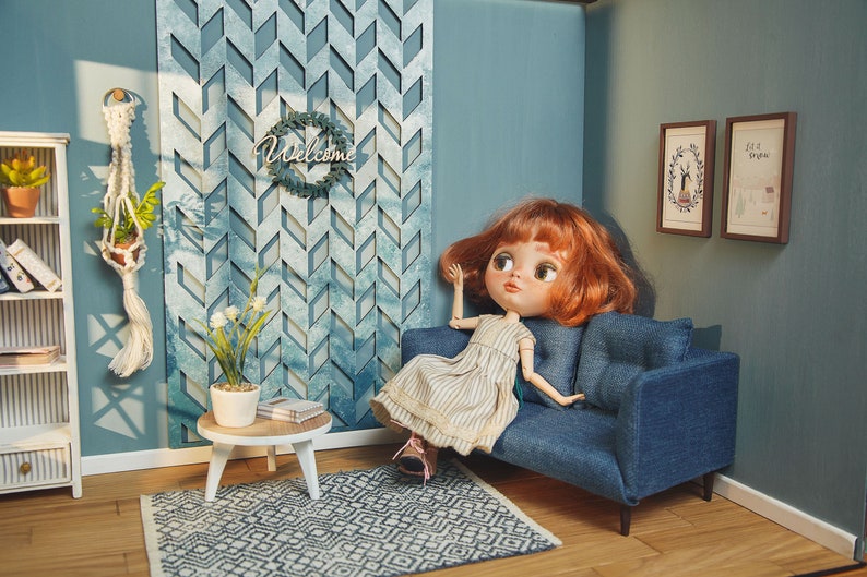 May include: A dollhouse living room with a blue couch, a white bookshelf, a small side table, and a doll with red hair sitting on the couch. The wall is blue and has a decorative wall panel with a geometric pattern. There is a welcome sign hanging on the wall.